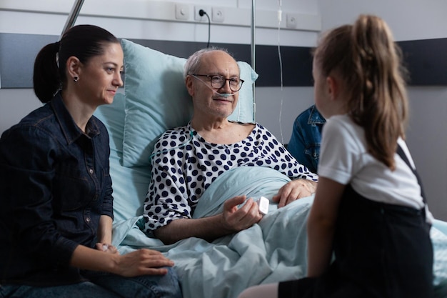 Uomo anziano sdraiato nel letto d'ospedale che chiacchiera con i membri della famiglia in visita. Bambina che parla con il nonno nella clinica medica degli anziani. Famiglia che vive con il vecchio che riposa nel letto del sanatorio.