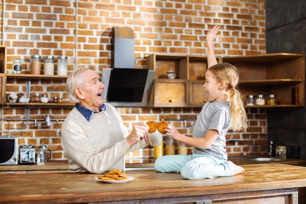 Uomo anziano sorridente allegro che assaggia la pasticceria fatta in casa con la sua nipote mentre levandosi in piedi in cucina