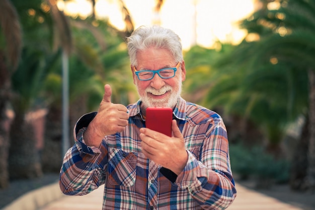 Uomo anziano sorridente che utilizza la tecnologia con lo smartphone per la videochiamata con la famiglia In piedi all'aperto in un parco pubblico al tramonto Felicità in faccia