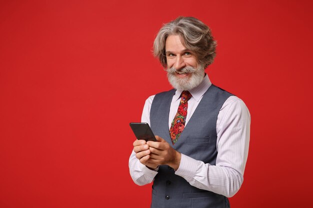 Uomo anziano sorridente con i baffi dai capelli grigi con la barba in classico gilet da camicia e cravatta colorata in posa isolata su sfondo rosso in studio. Concetto di stile di vita delle persone. Mock up spazio di copia. Tenendo il cellulare.