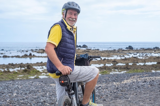 Uomo anziano sorridente del ciclista attivo che indossa il casco in piedi lungo la spiaggia del mare sulla sua bicicletta elettrica guardando la fotocamera godendo di uno stile di vita sano