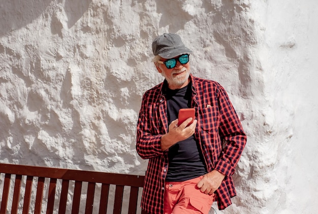 Uomo anziano sorridente in camicia rossa a scacchi in piedi all'aperto contro un muro mentre utilizza il telefono cellulare