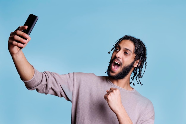 Uomo arabo eccitato che prende selfie sullo smartphone mentre mostra il gesto del vincitore con il pugno chiuso. Giovane persona allegra che posa per la foto del telefono cellulare con l'espressione facciale felice