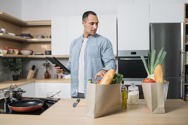 Uomo arabo felice che cucina cibo sano con ricetta che guarda su sacchetti di carta con verdure