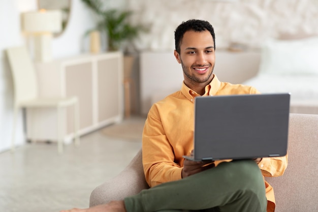 Uomo arabo felice che utilizza il computer portatile che si siede sul sofà a casa