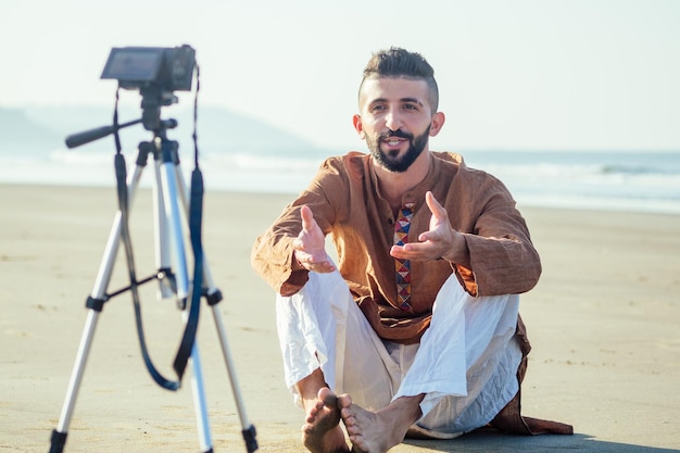 Uomo arabo seduto sulla sabbia e parlare di video treppiede in spiaggia Concetto di creatore di contenuti lifestyle per videografi di viaggio