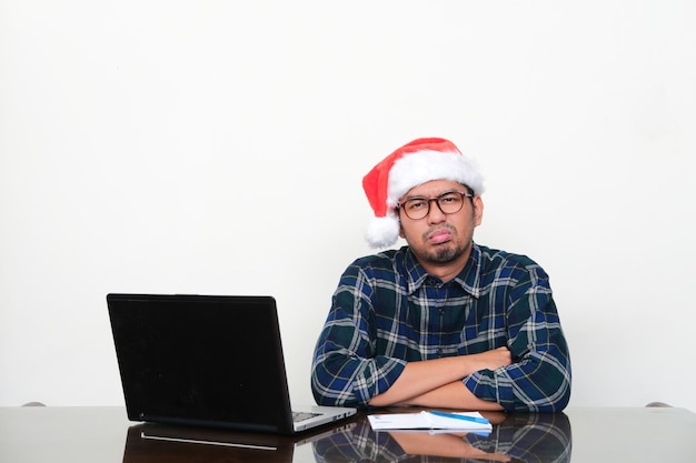 Uomo asiatico adulto che indossa cappello di Natale seduto nel suo tavolo di lavoro con espressione pigra