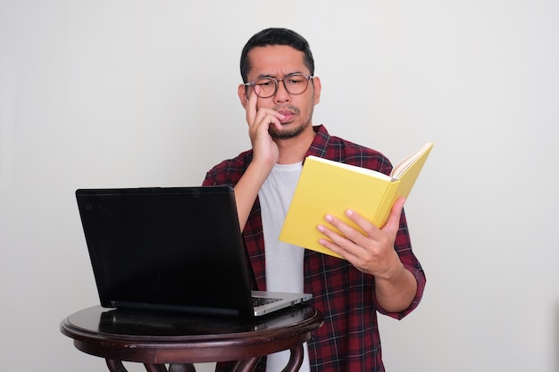 Uomo asiatico adulto che legge un libro davanti al computer portatile con un'espressione seria del viso