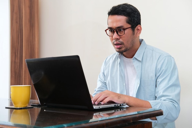 Uomo asiatico adulto che mostra il viso scioccato quando guarda al suo laptop