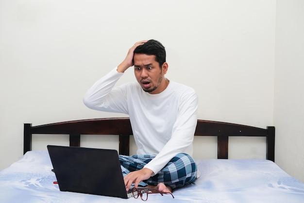 Uomo asiatico adulto che mostra un'espressione scioccata mentre guarda al computer portatile nel suo letto