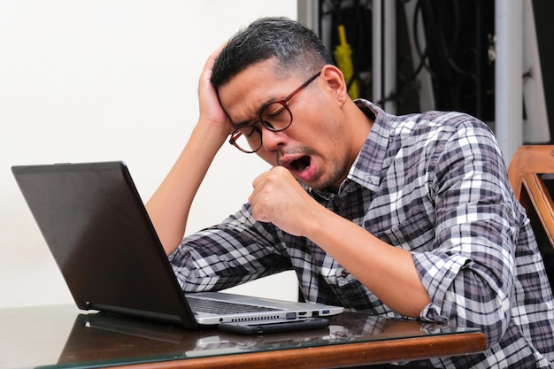 Uomo asiatico adulto che sbadiglia quando lavora usando il portatile mostrando stanchezza