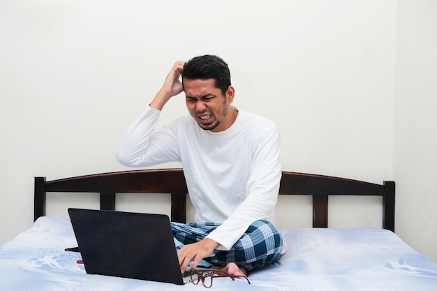 Uomo asiatico adulto che sembra stressato mentre lavora utilizzando il laptop nel suo letto