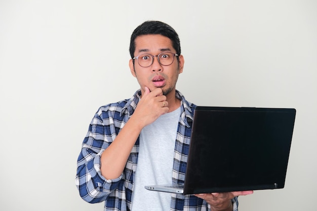 Uomo asiatico adulto che si tocca il mento mostrando un gesto dubbioso mentre tiene un laptop