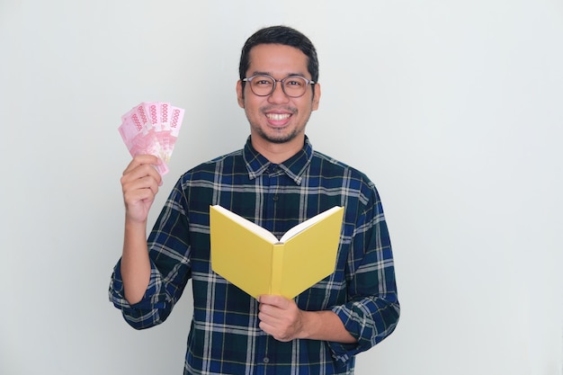 Uomo asiatico adulto che sorride felice alla macchina fotografica mentre tiene un libro e soldi