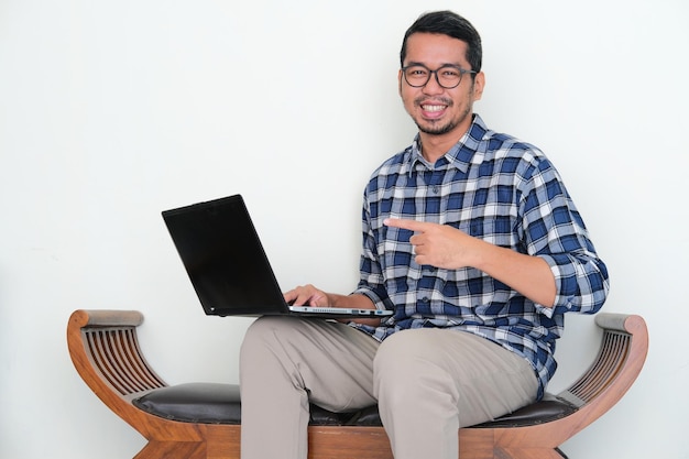 Uomo asiatico adulto seduto su una panchina che sorride fiducioso mentre indica il suo laptop
