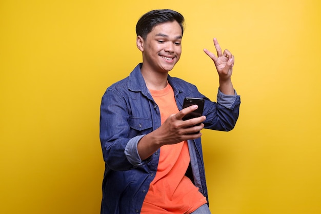 Uomo asiatico allegro che mostra il dito del segno v e sorride felice mentre si fa un selfie su uno sfondo giallo
