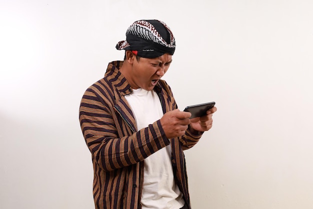 Uomo asiatico arrabbiato e confuso in costume giavanese in piedi mentre usa uno smartphone