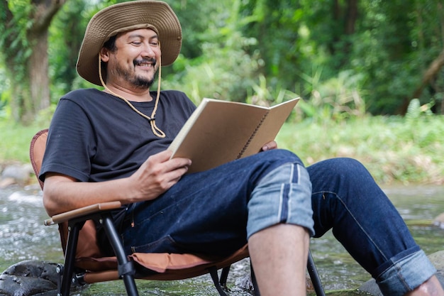 Uomo asiatico barbuto felice seduto da solo su una sedia vicino a un ruscello in campagna e godendosi il paesaggio della foresta mentre legge un libro Leggere o studiare un concetto