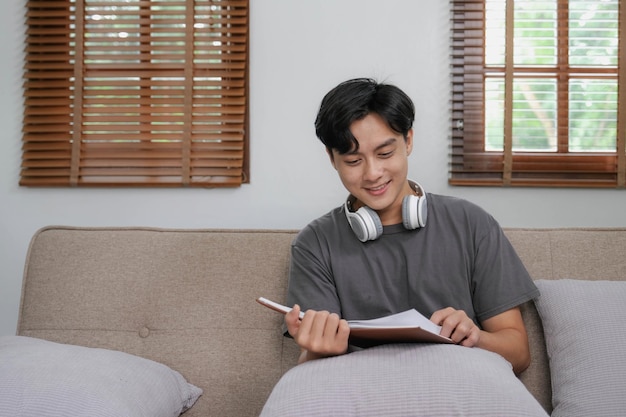 Uomo asiatico bello che si rilassa sul divano di casa con un libro e le cuffie