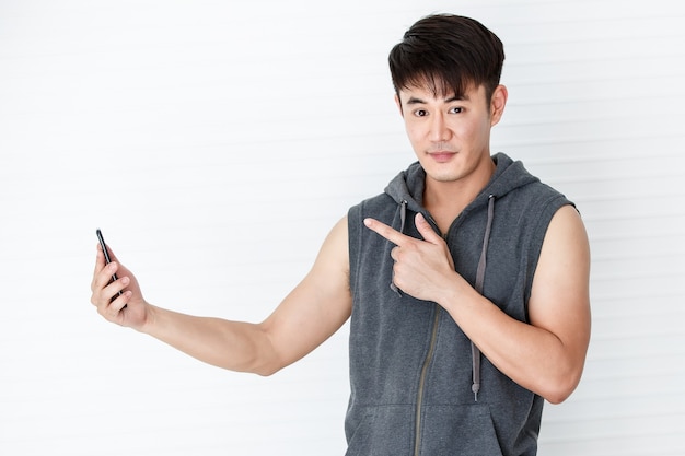 Uomo asiatico bello in forma sorridente che tiene la mano smartphone prendendo un gesto felice selfie indossando t-shirt senza maniche abbigliamento sportivo grigio in sfondo bianco.