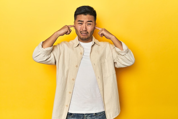 Uomo asiatico casuale con camicia aperta T-shirt bianca su studio giallo che copre le orecchie con le dita sottolineate