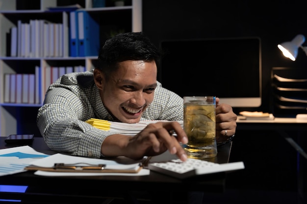 Uomo asiatico che fa gli straordinari a bere birra mentre utilizza la calcolatrice in ufficio di notte