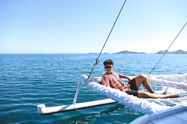 Uomo asiatico che indossa occhiali da sole che si rilassano sulla rete del catamarano godendosi l'ora legale