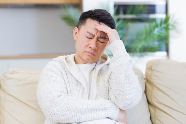 uomo asiatico che si tiene la testa con un forte mal di testa a casa in una stanza sul divano