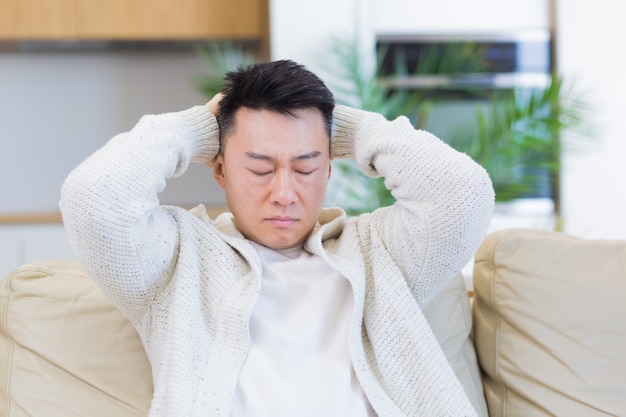 uomo asiatico che si tiene la testa con un forte mal di testa a casa in una stanza sul divano