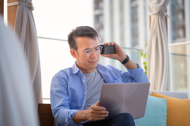 Uomo asiatico che utilizza smartphone e laptop seduto sul divano di casa Uomo che lavora da un laptop sulla terrazza