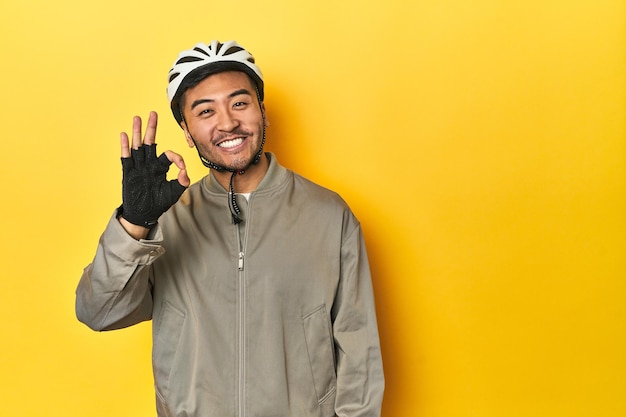 Uomo asiatico con casco da bicicletta e guanti sullo sfondo giallo dello studio allegro e sicuro mostrando un gesto ok