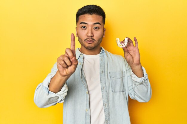 Uomo asiatico con correttore dentale invisibile sullo sfondo giallo dello studio che mostra il numero uno con il dito