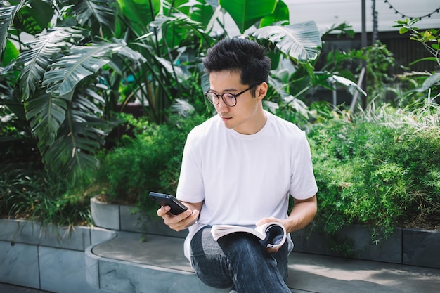 Uomo asiatico con libro utilizzando smartphone in giardino
