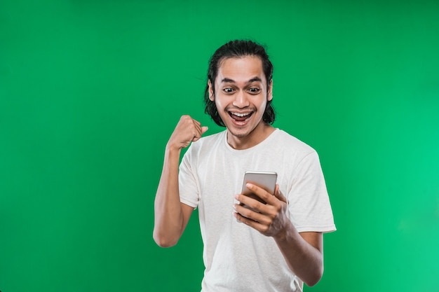 Uomo asiatico con lunghi capelli neri che tiene e guarda handphone con espressione di felicità mentre alza una mano e fa un pugno, isolato su sfondo verde