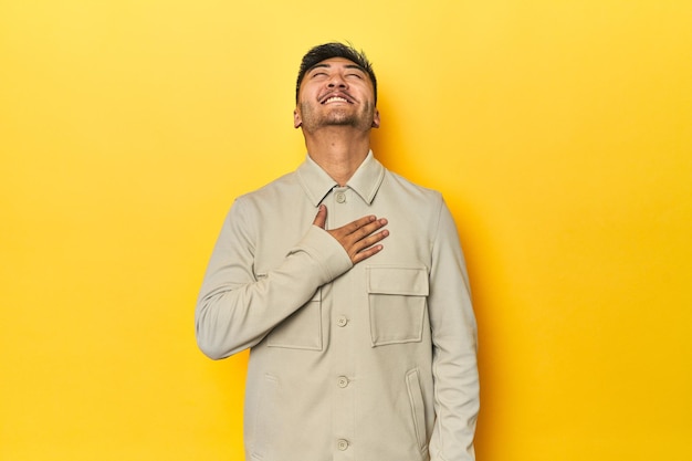 Uomo asiatico con una elegante camicia grigia su uno studio giallo ride ad alta voce tenendo la mano sul petto