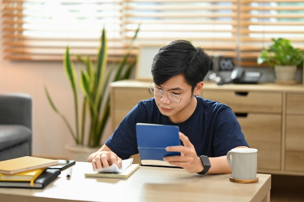 Uomo asiatico concentrato che calcola le finanze domestiche o le tasse sulla calcolatrice seduto in un soggiorno luminoso