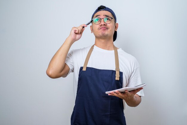 Uomo asiatico del barista in grembiule blu che prende la scrittura dell'ordine sulla lista del libro del menu che pensa con la penna sulla testa