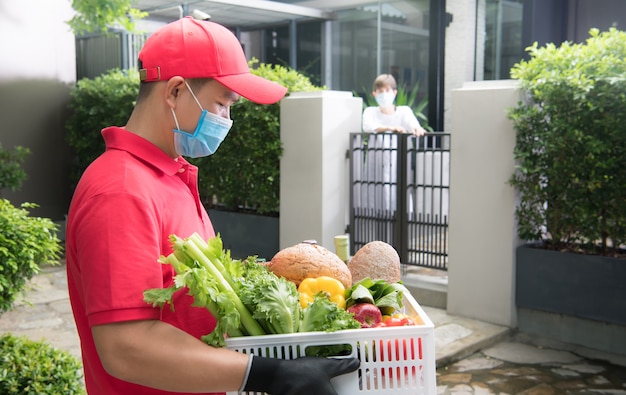 Uomo asiatico di consegna che indossa la maschera e guanti in uniforme rossa che trasporta la scatola di generi alimentari