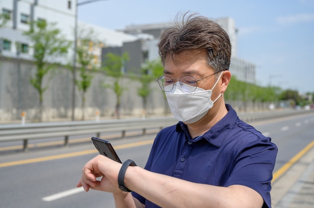 Uomo asiatico di mezza età che indossa una maschera facciale e guarda uno smartphone nel vialetto.