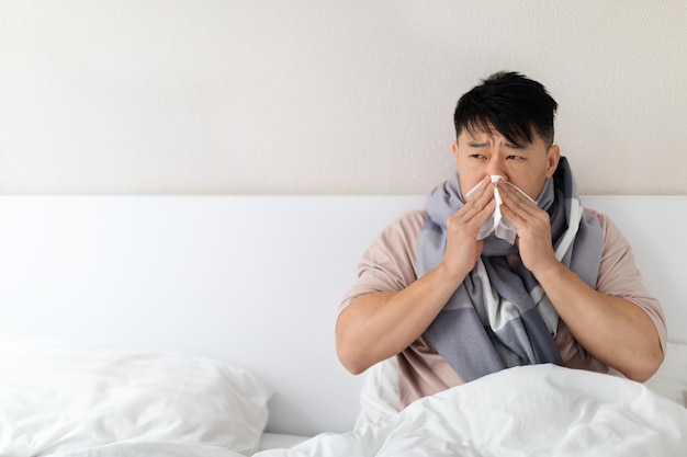 Uomo asiatico di mezza età malato seduto a letto che starnutisce