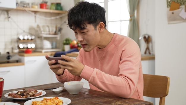 uomo asiatico dipendente che gioca con il telefono cellulare mentre pranza nella sala da pranzo di casa. posa le bacchette e usa il telefono sentendosi eccitato