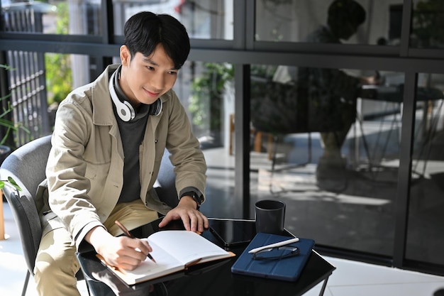 Uomo asiatico felice che fa elenchi sul suo libro di pianificatore personale che lavora a distanza presso la caffetteria