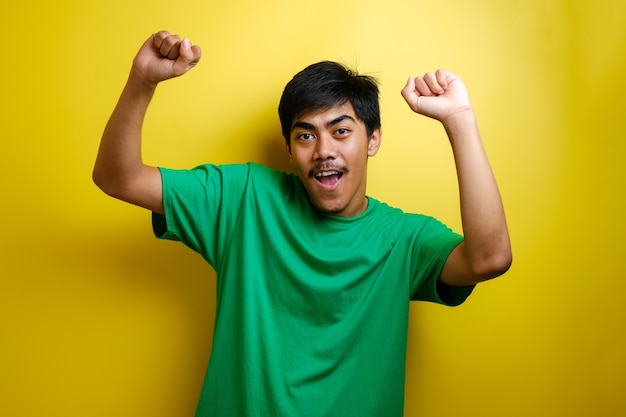 Uomo asiatico in maglietta verde che sorride e balla felicemente, gioioso che esprime celebrando la buona notizia vittoria vincendo il gesto di successo su sfondo giallo