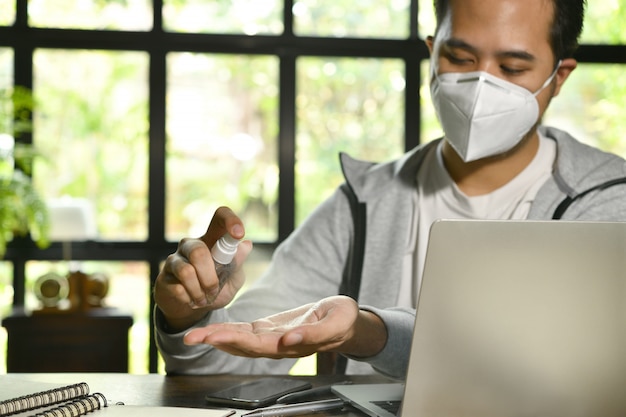 Uomo asiatico in quarantena e distanza sociale che indossa una maschera chirurgica e si pulisce le mani con disinfettante in gel alcolico mentre lavora da casa durante la pandemia di Coronavirus Covid-19