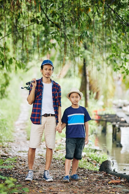 Uomo asiatico maturo che tiene la mano di suo figlio quando stanno camminando verso il lago con l'asta