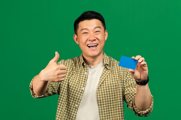 Uomo asiatico maturo felice che tiene la carta di credito e che mostra il pollice in su consigliando il cliente della banca maschio