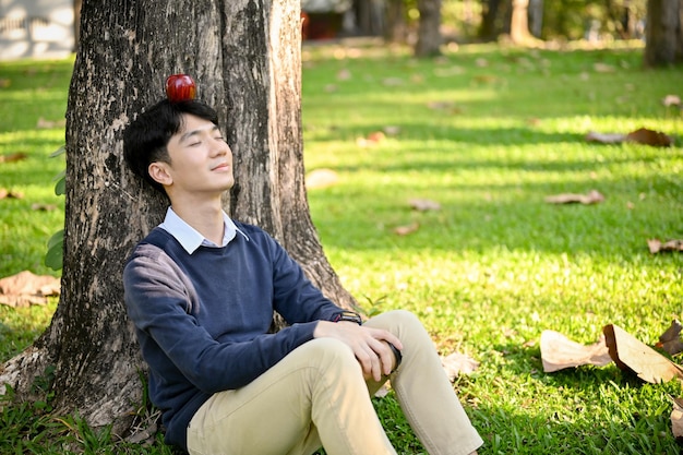 Uomo asiatico rilassato seduto sotto gli occhi dell'albero chiuso agghiacciante nel bellissimo parco verde