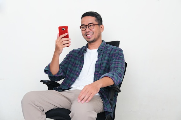 Uomo asiatico seduto su una sedia da ufficio con un'espressione felice quando guarda al suo telefono