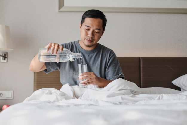 Uomo asiatico seduto sul letto che versa acqua minerale da una bottiglia a un bicchiere