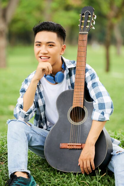 Uomo asiatico seduto sull'erba verde con la chitarra e sorride alla macchina fotografica all'aperto